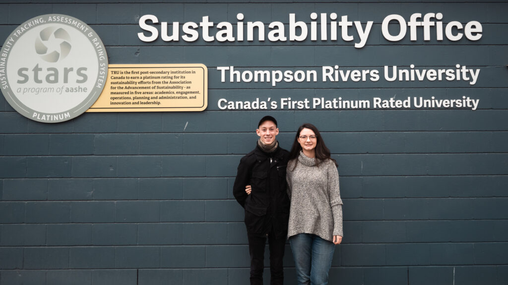A young man wearing dark clothing stands to the left of a young lady wearing a light grey sweater. The individuals are standing adjacent to a sign for the TRU Sustainability Office