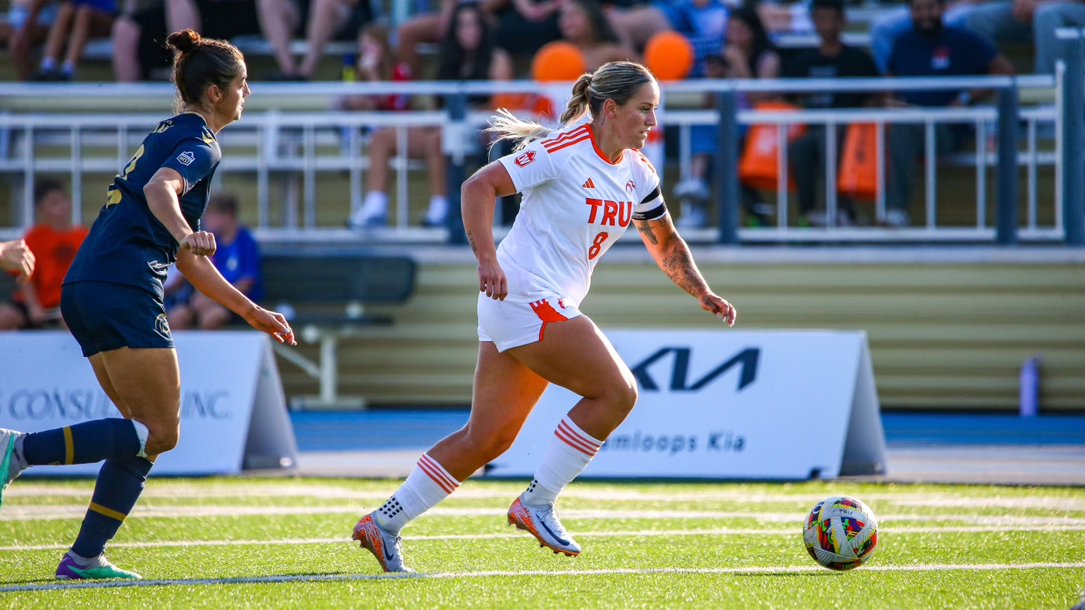 A woman dressed primarily in black chases another woman wearing primarily white on a soccer field. Both are chasing after a soccer ball.