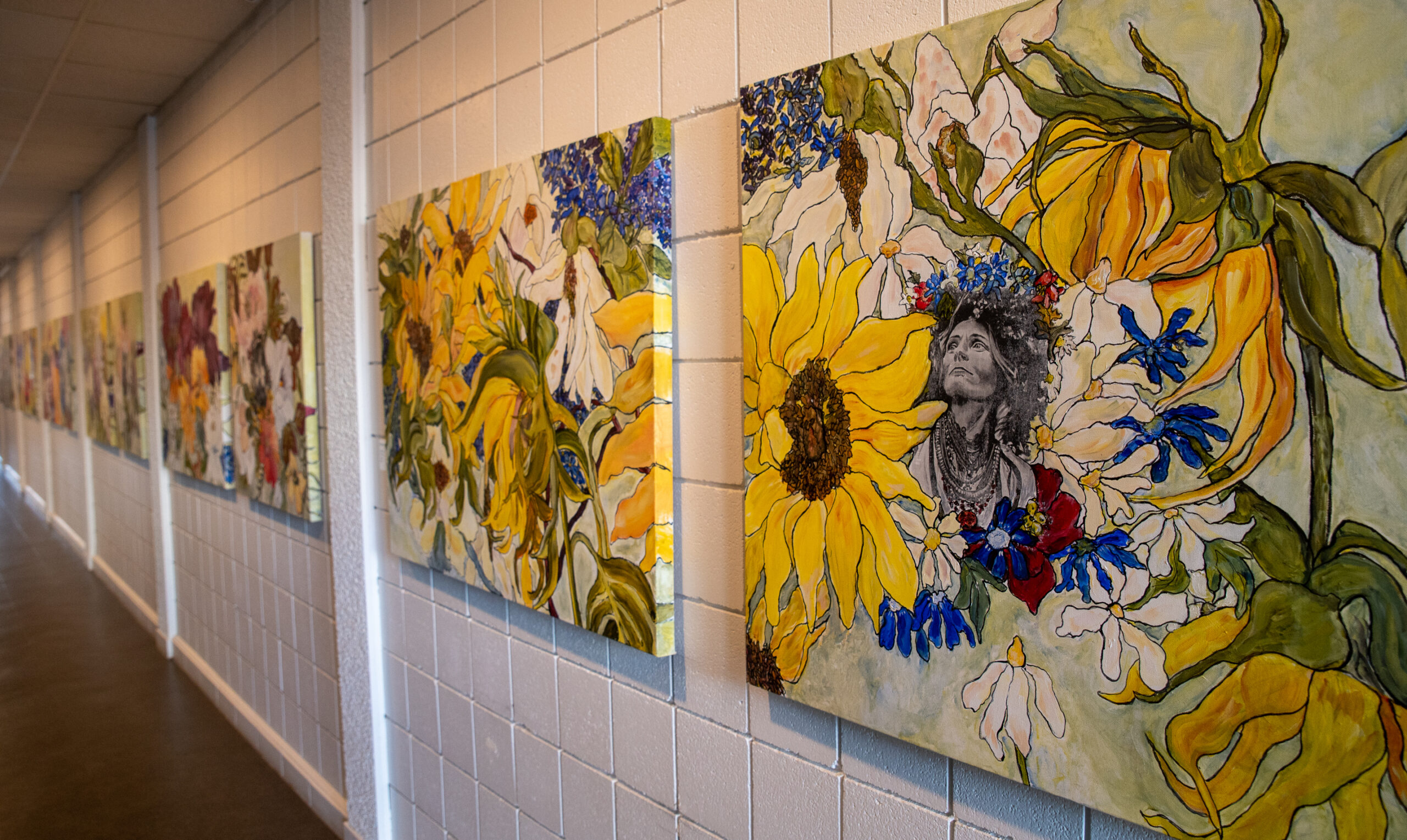 A row of paintings hang on a wall in a well lit hallway. Many of the paintings share the same yellow and blue palettes.