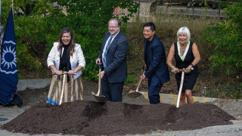 A woman in white holding a petse and a long skirt stands next two men wearing dark suits, and a second woman in black, standing atop a dirt pile for a groundbreaking ceremony