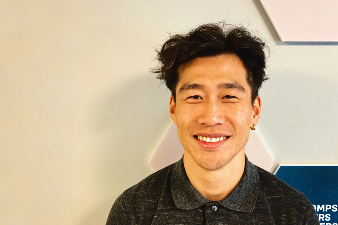 A man with fluffy black hair is seen smiling, standing against an off-white background. Photo was taken at Thompson Rivers University in Kamloops, BC, September 2024.