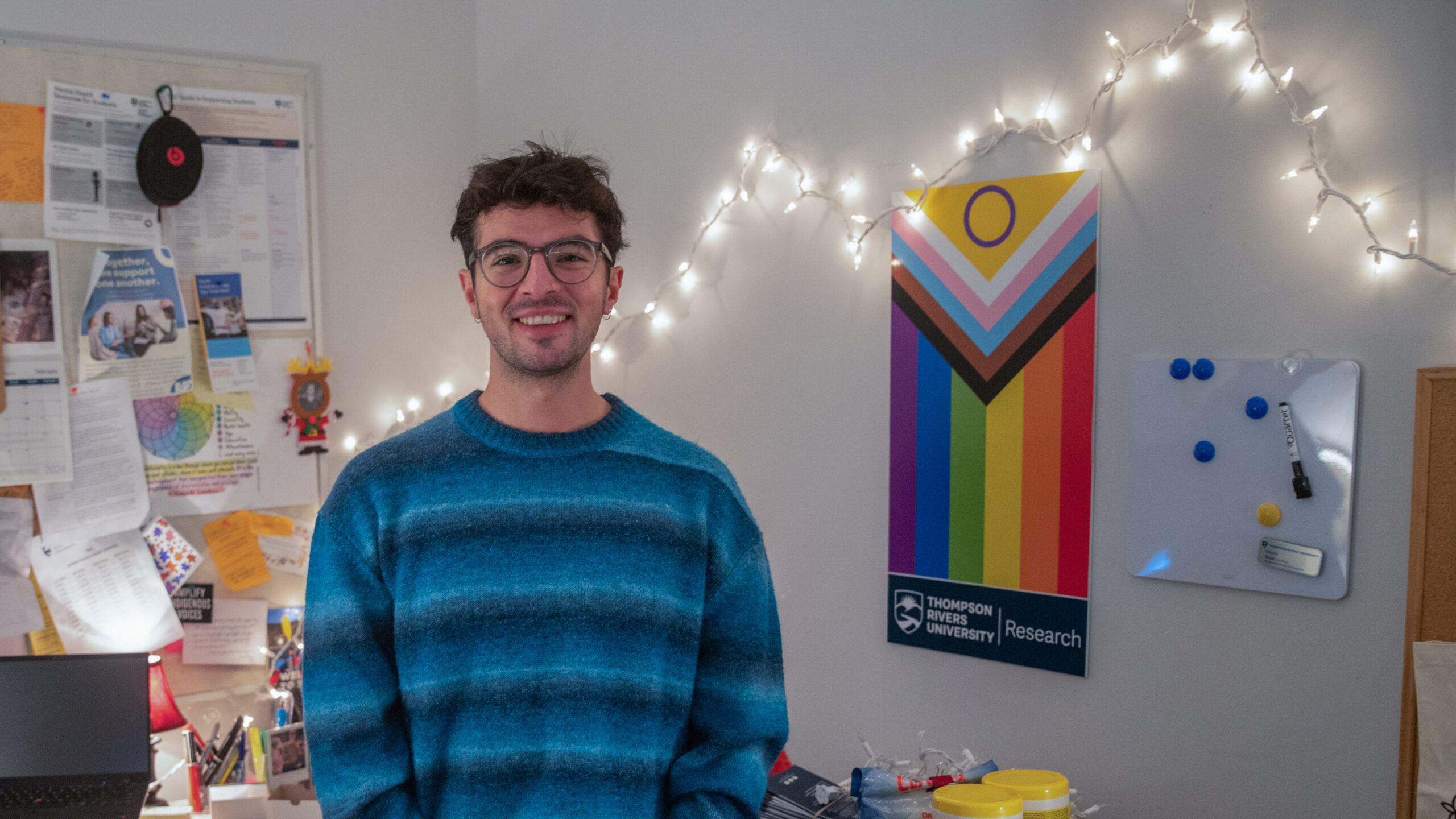 A person with brown hair and glasses poses for a portrait in an office with a Pride flag and string lights in the background.